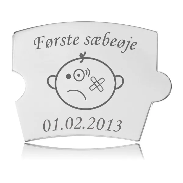 Memozz Spin brik i poleret rustfrit stål med teksten "Første sæbeøje" og tegning af barneansigt med forbinding