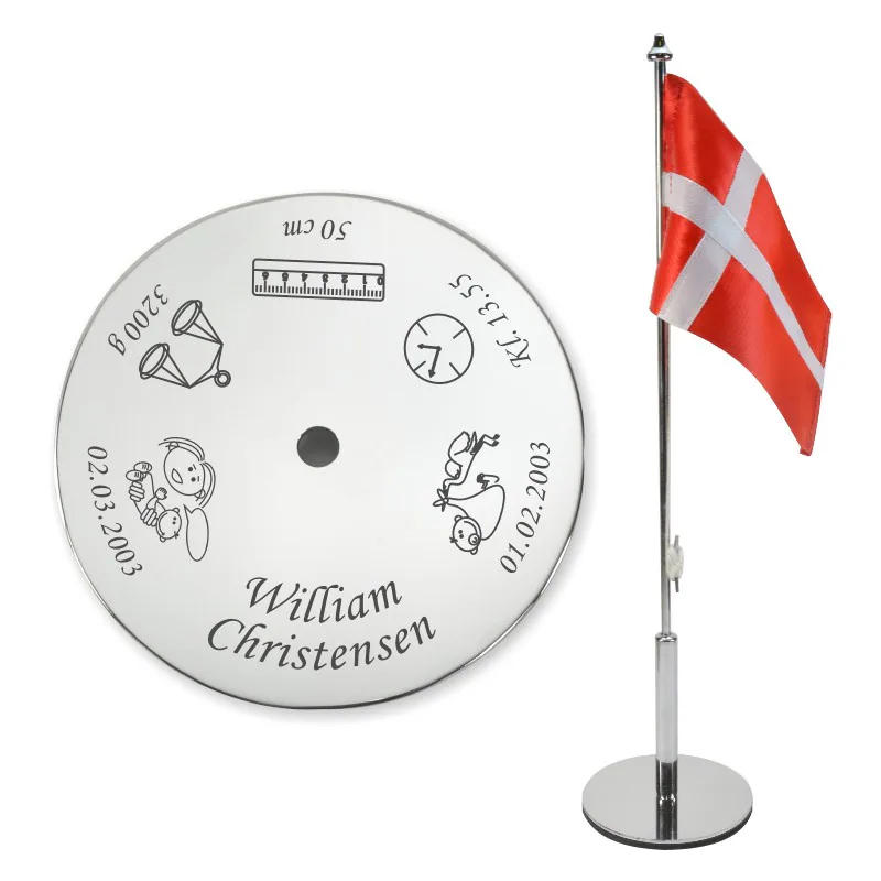 Navngivningsflag i poleret stål med personlig gravering af navn og fødselsdata, vist ved siden af det håndsyede danske flag.