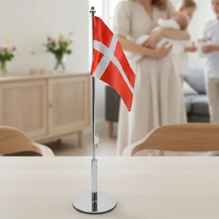Et dansk bordflag på en metalstang stående på et lyst træbord med en sløret familie i baggrunden til en festlig begivenhed.