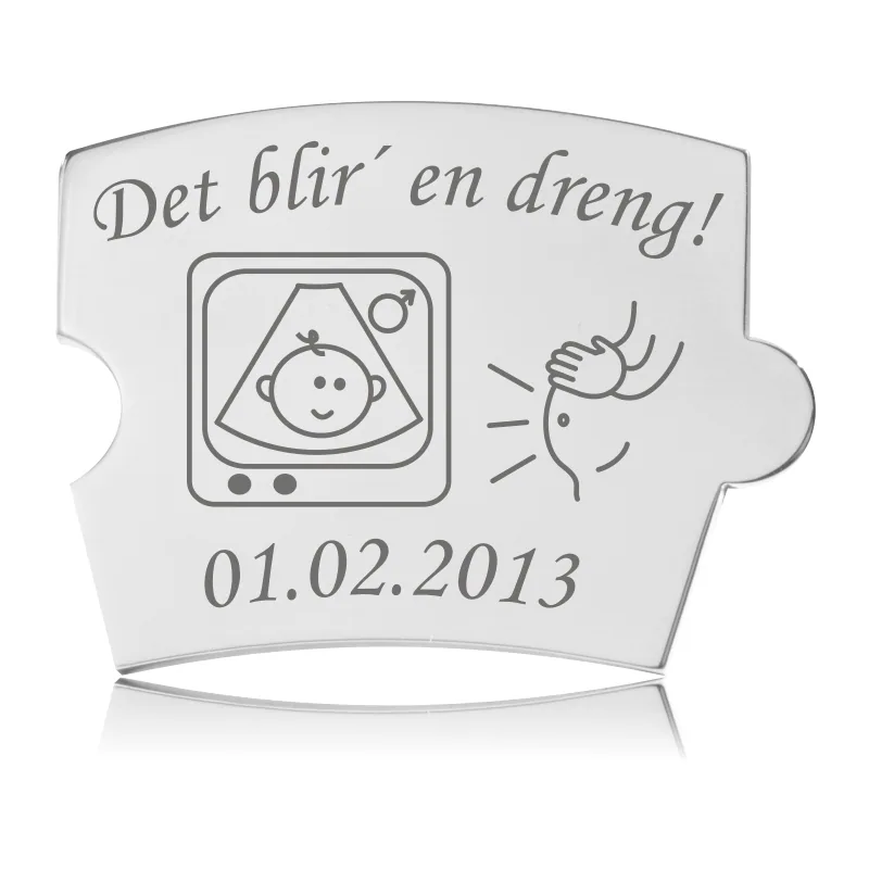 Memozz Spin brik i håndpoleret rustfrit stål med teksten "Det blir' en dreng!", sonogram-ikon og hænder på gravid mave.