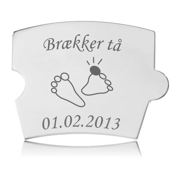 Rektangulær Memozz Spin brik i håndpoleret rustfrit stål med gravering af teksten "Brækker tå", to små fødder med en rød prik på en tå, samt datoen "01.02.2013".